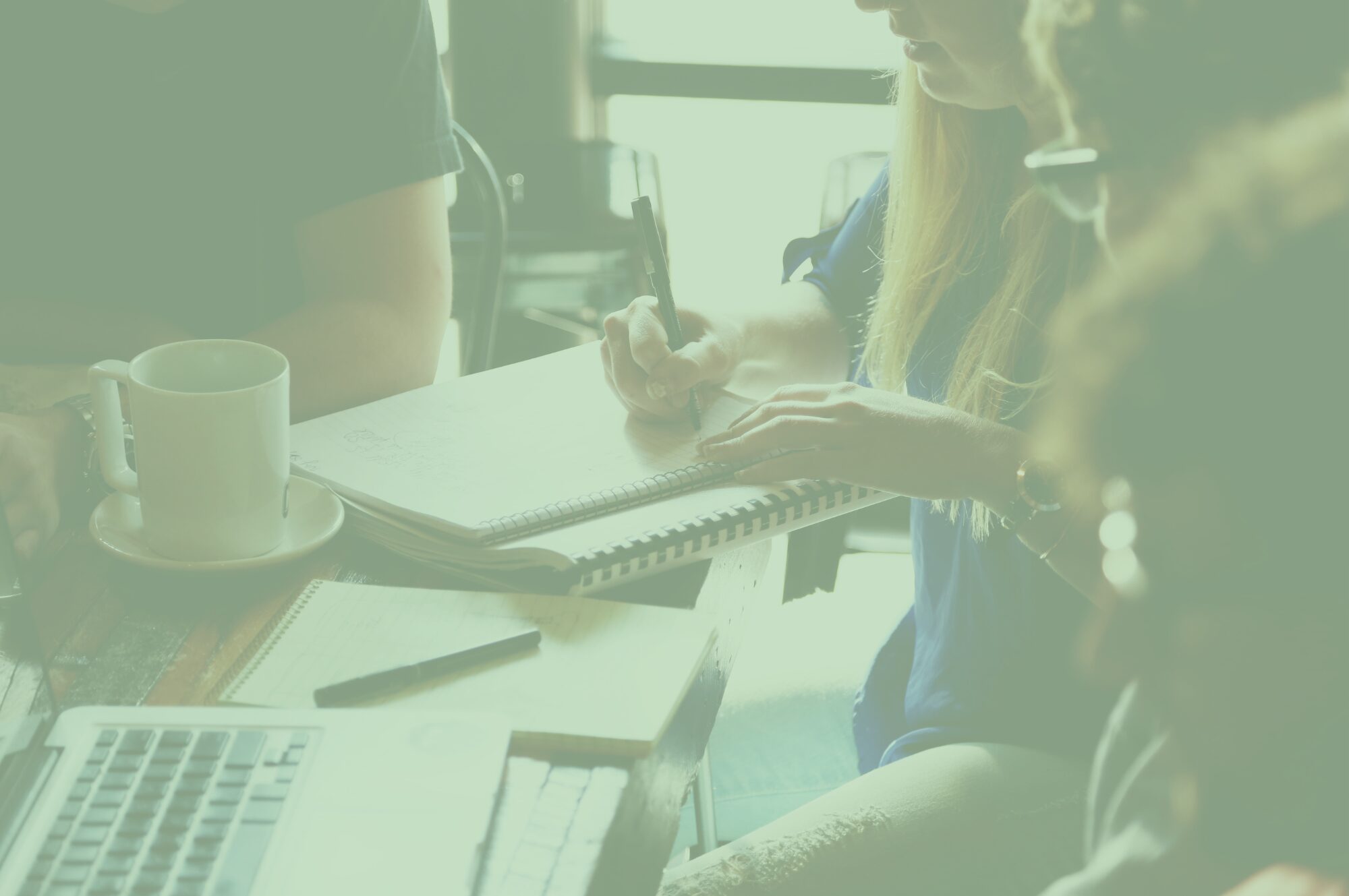 Three people sit round a table making notes, with laptops, pens, notepads and coffee on the table.