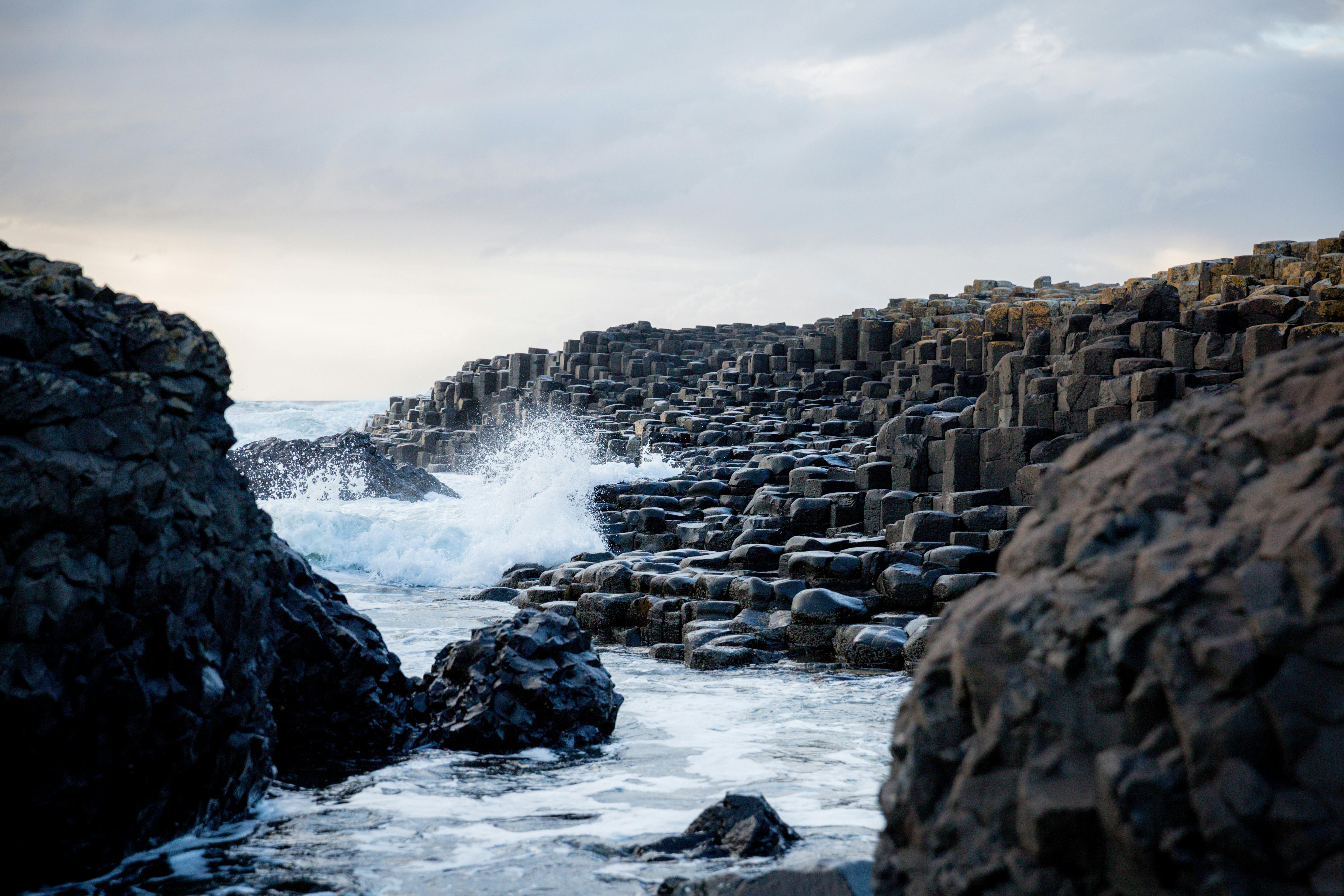 The hexagonal basalt columns of the Giant's Causeway in Northern Ireland extend into the sea as white waves crash against the rocks under a dark, cloudy sky.